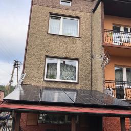 Sławomir Wasilik Voltum Kompleksowe Usługi Elektryczne - Panele fotowoltaiczne na dachu budynku z widoczną drabiną opartą o ścianę, słup elektryczny i antenę telewizyjną na elewacji.