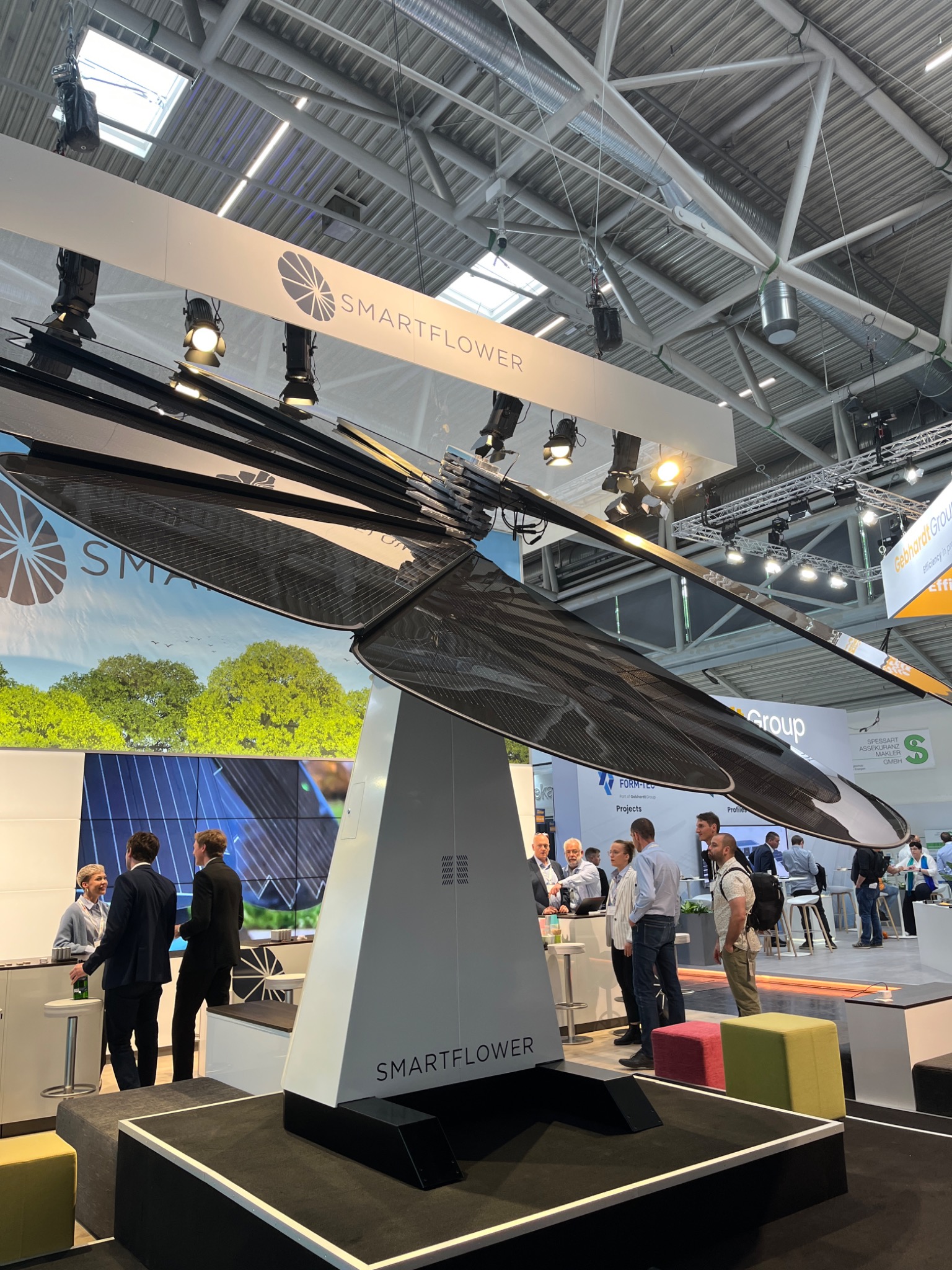 Nowoczesna instalacja fotowoltaiczna Smartflower na targach, otoczona przez osoby oglądające. Unikalny design przypominający kwiat, z logo i oświetleniem.
