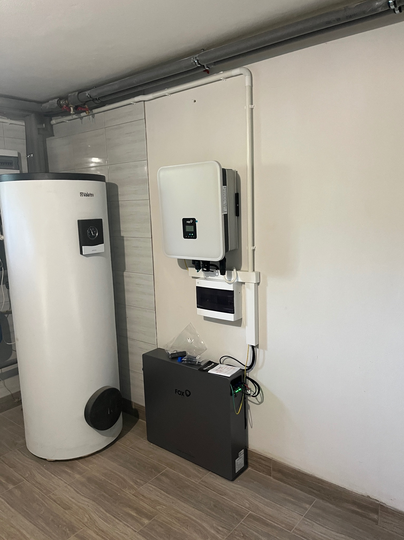 Biały inwerter fotowoltaiczny H2O, akumulator Fox-Ess i zbiornik Valami zamontowane na ścianie w pomieszczeniu z brązową podłogą imitującą drewno.