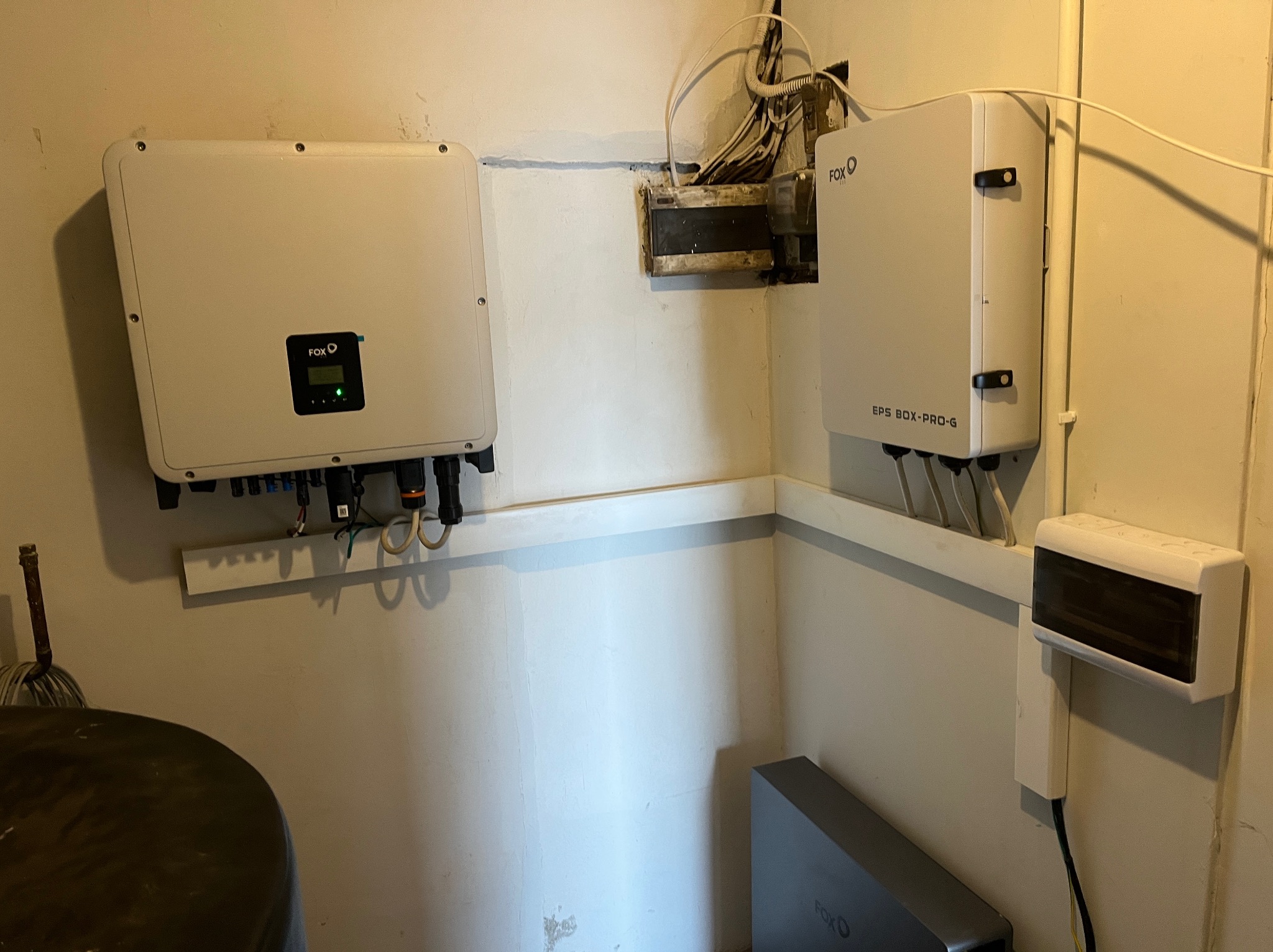 Zamontowany inwerter FOX i skrzynka EPS BOX-PRO-G na ścianie w pomieszczeniu. Widoczne kable i instalacja elektryczna. Elementy systemu fotowoltaicznego w rogu pomieszczenia.