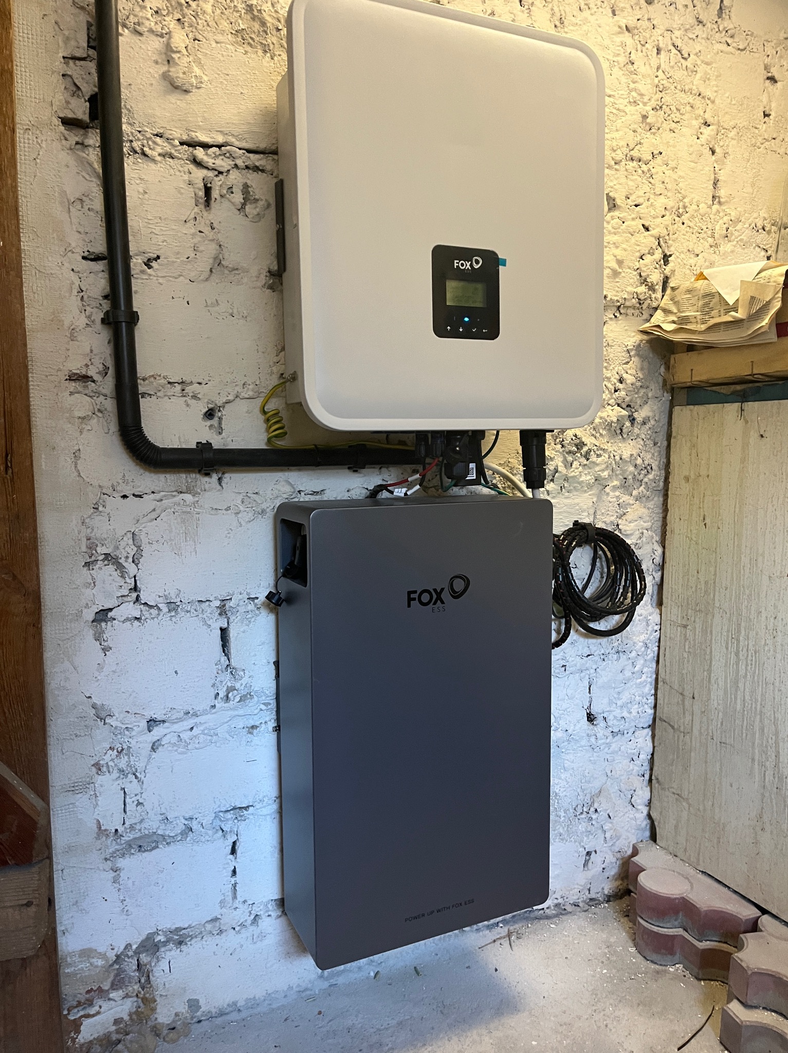 Instalacja falownika FoxESS i modułu bateryjnego w kotłowni we Wrocławiu. Biała, nierówna ściana z cegły, czarne kable. Elementy systemu fotowoltaicznego.