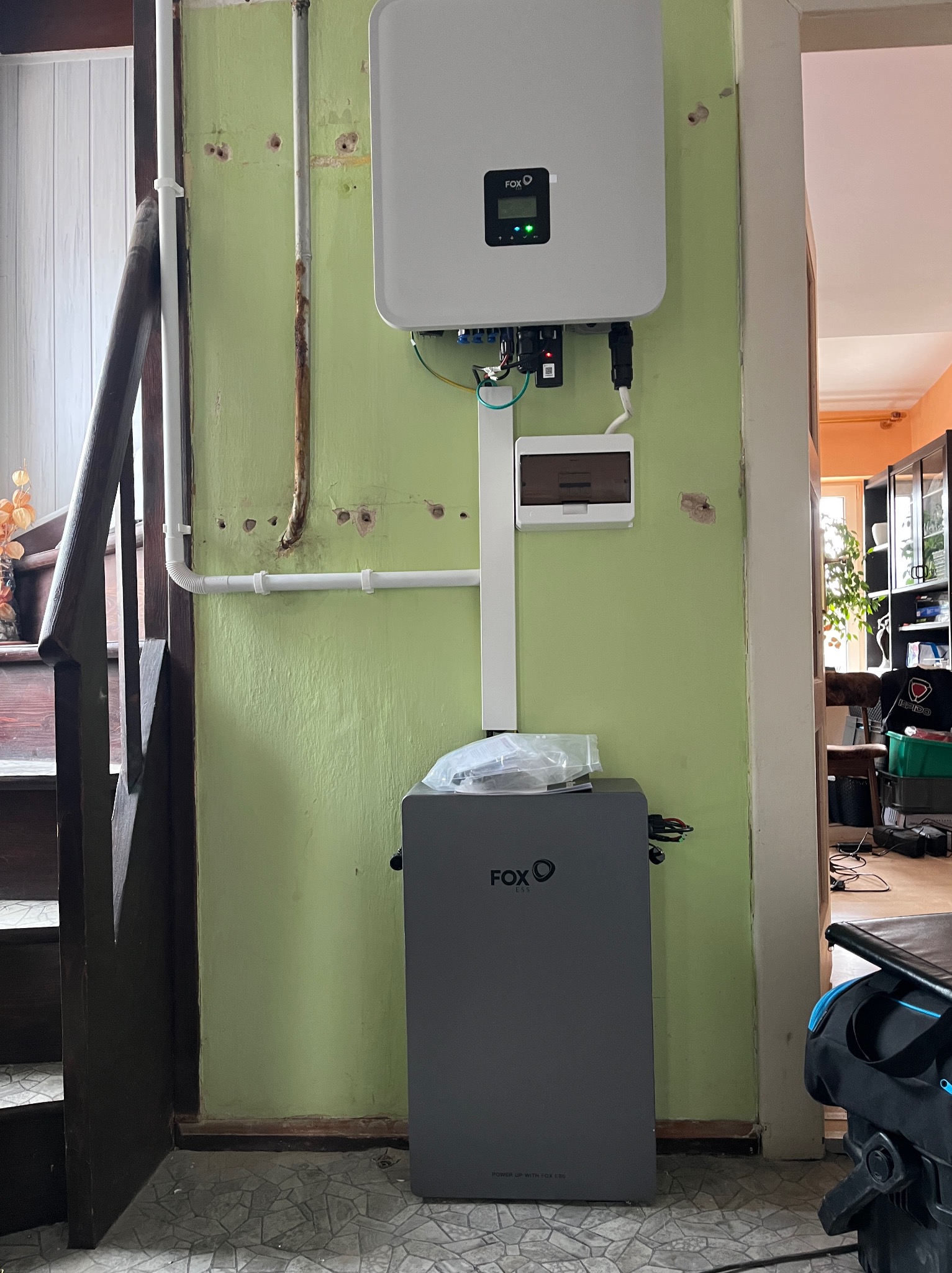 Szary akumulator i biały inwerter FOX ESS zamontowane na zielonej ścianie w korytarzu domu, obok schodów. Widoczne przewody i skrzynka elektryczna.