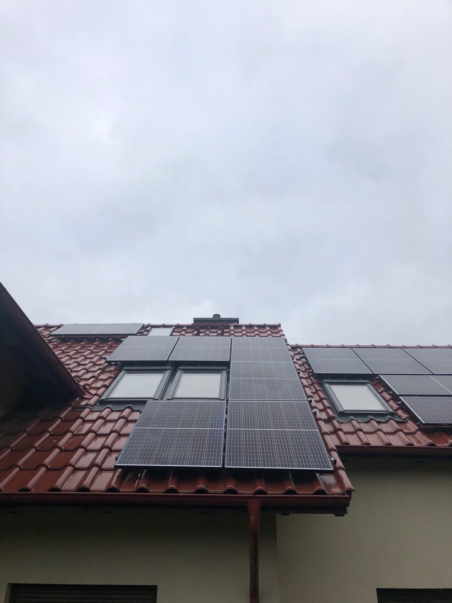 Panele fotowoltaiczne zamontowane na dachu z czerwonej dachówki, widoczne okna dachowe, pochmurne niebo w tle.