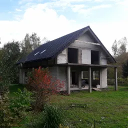 Surowy stan dwukondygnacyjnego domu z czarnym dachem w trakcie budowy, widoczne bloczki betonowe, otoczenie zielone, drzewa i częściowo żółty samochód dostawczy w tle.