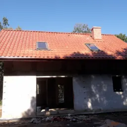 Surowy stan domu jednorodzinnego z czerwonym dachem, widoczne okna dachowe i komin z cegły, ściany z betonu komórkowego, teren wokół domu w trakcie prac ziemnych.