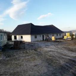 Budowa domu jednorodzinnego w stanie surowym otwartym, widoczny fundament, ściany z bloczków i dachówka, obok przyczepa kempingowa i materiały budowlane.