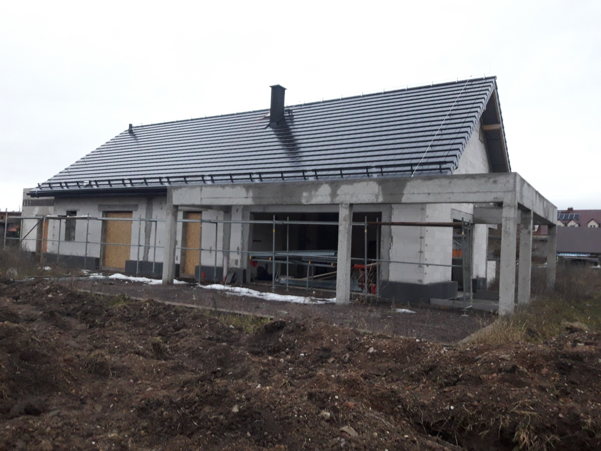 Surowy stan domu jednorodzinnego w trakcie budowy, widoczne ściany z betonu komórkowego, czarna dachówka, teren wokół domu z ziemią i resztkami śniegu.