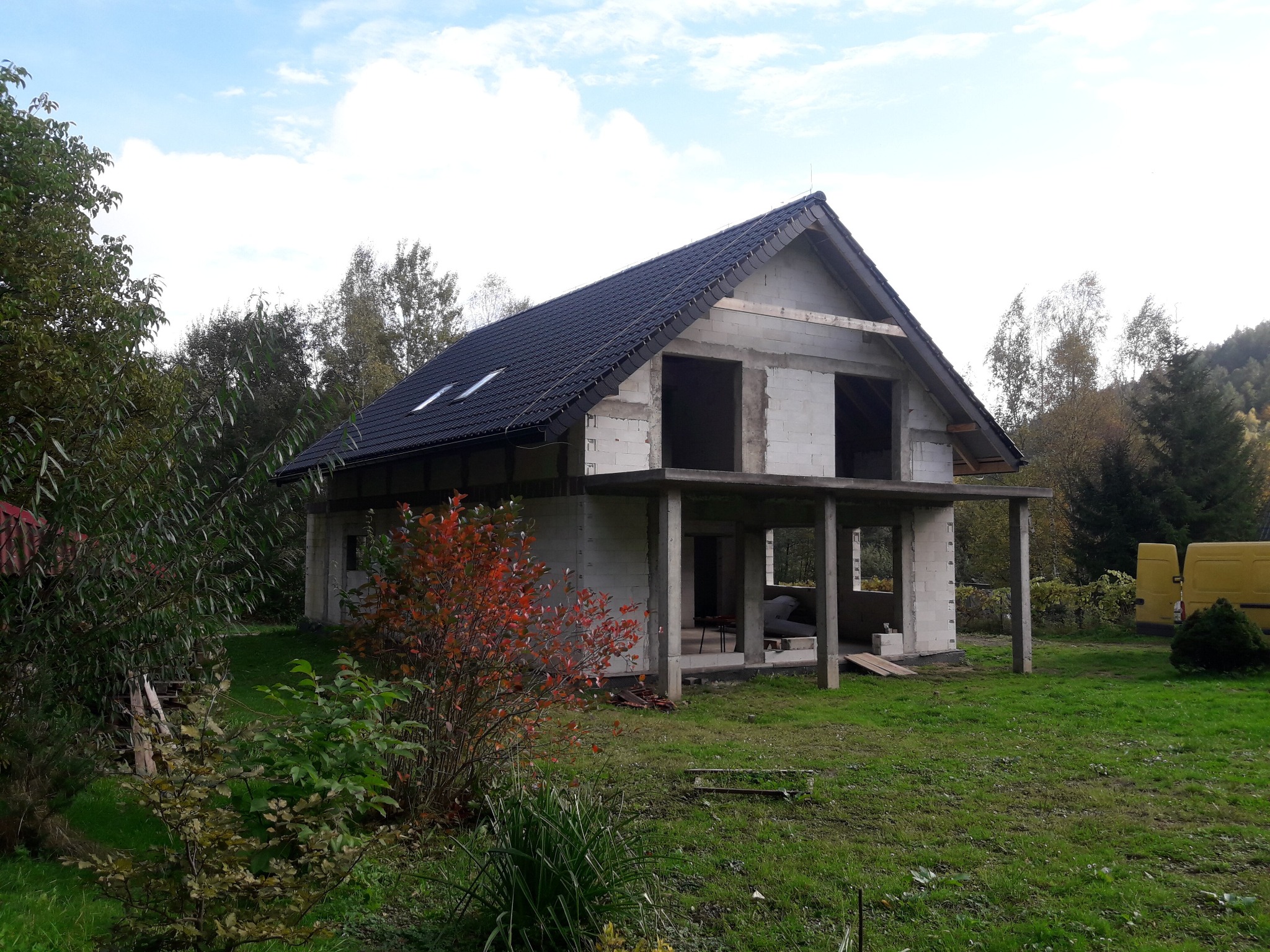 Surowy stan dwukondygnacyjnego domu z czarnym dachem w trakcie budowy, widoczne bloczki betonowe, otoczenie zielone, drzewa i częściowo żółty samochód dostawczy w tle.