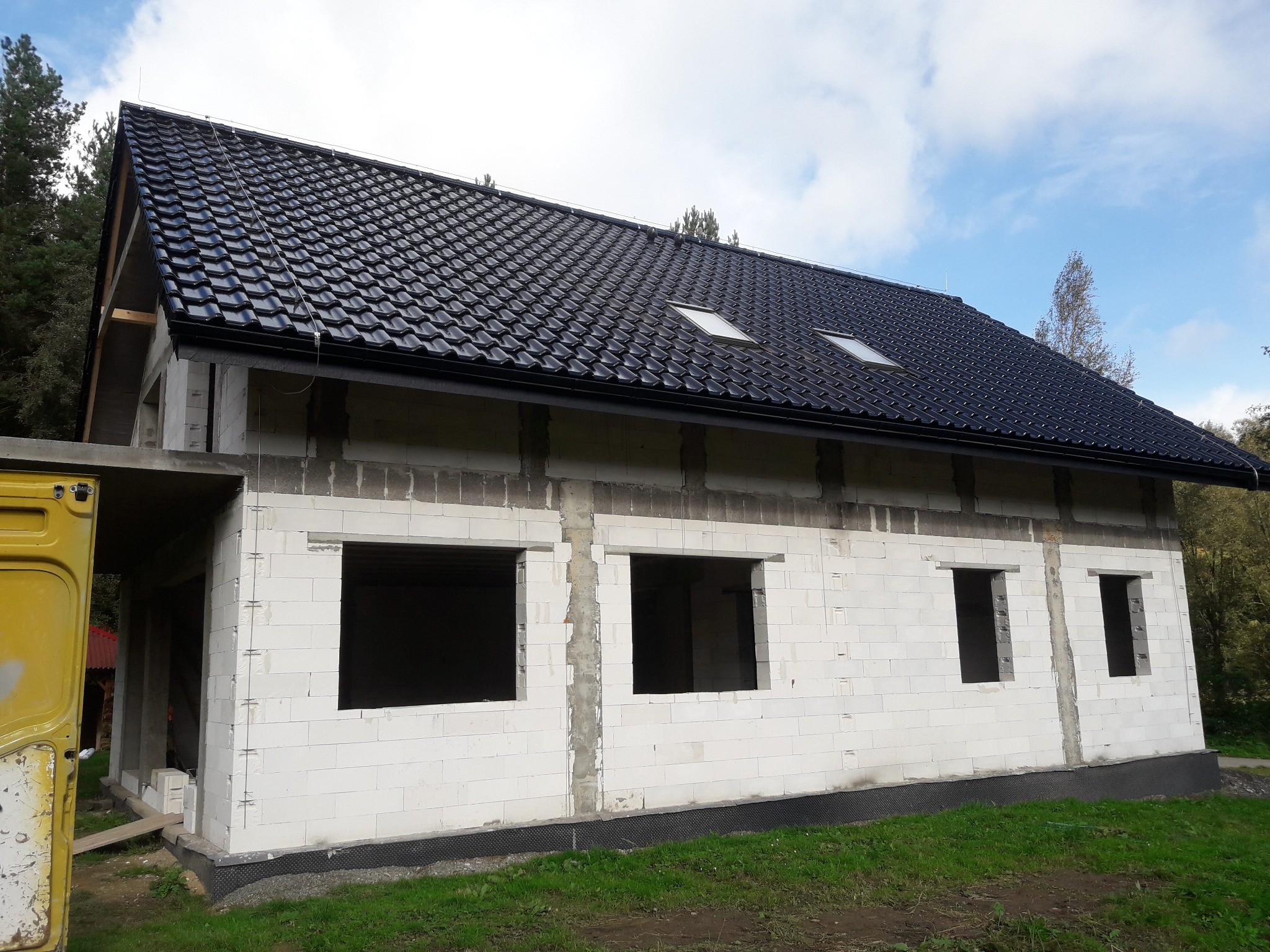 Wznoszenie ścian domu z białych bloczków, widoczna czarna hydroizolacja fundamentów i dach z dwoma oknami dachowymi, żółty samochód dostawczy w tle.