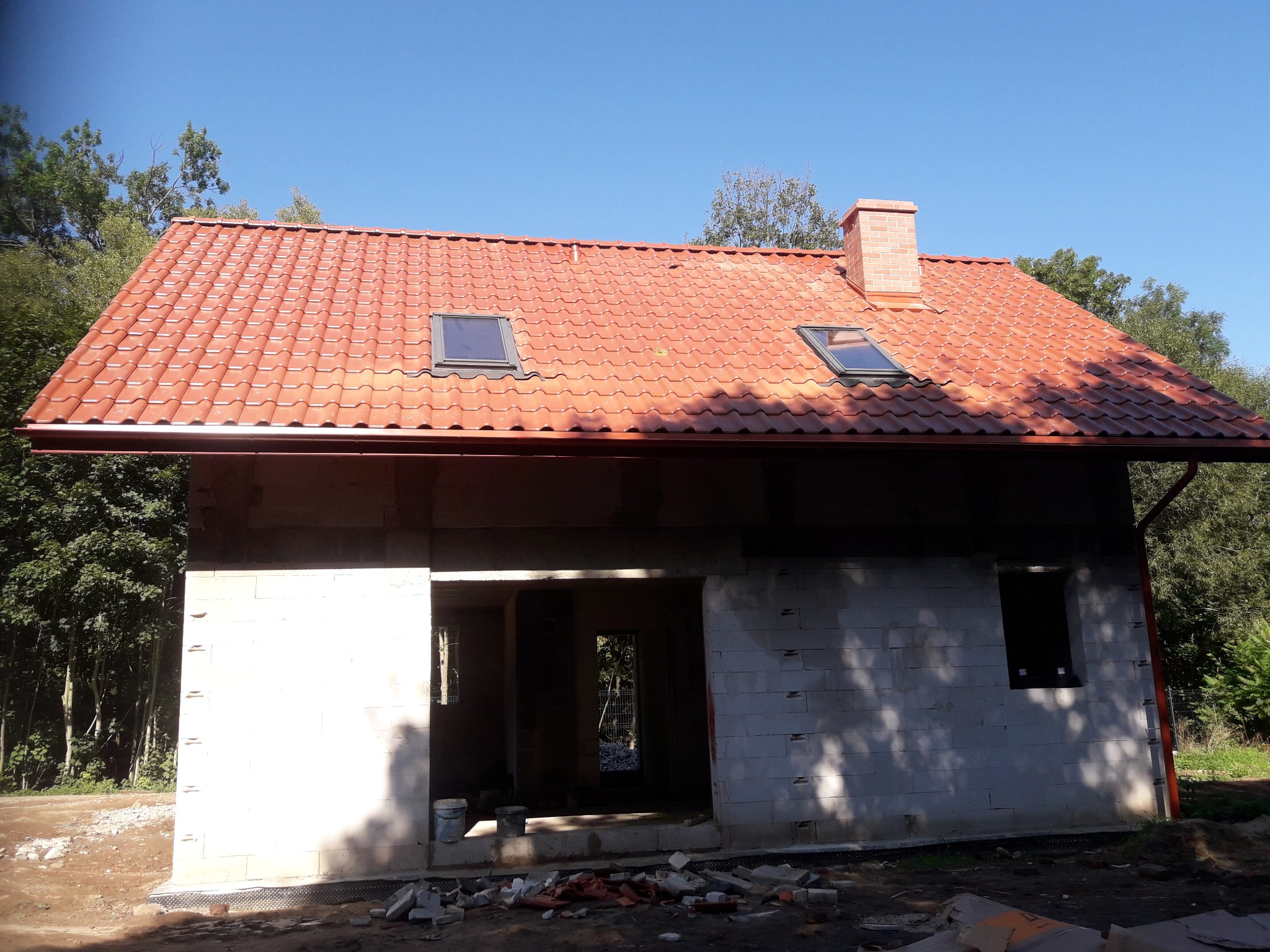 Surowy stan domu jednorodzinnego z czerwonym dachem, widoczne okna dachowe i komin z cegły, ściany z betonu komórkowego, teren wokół domu w trakcie prac ziemnych.