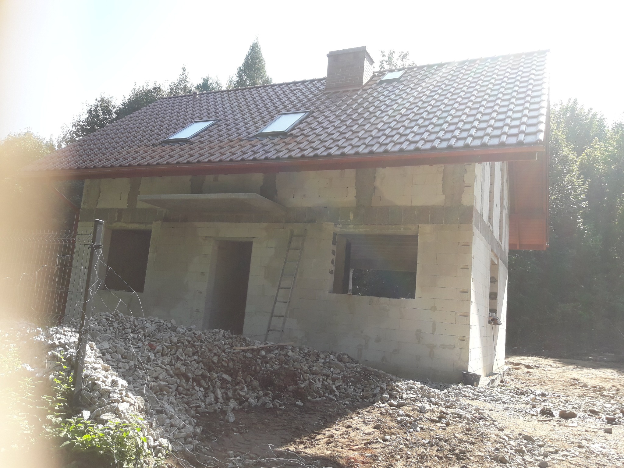 Niedokończony dwupiętrowy dom w stanie surowym otwartym z brązową dachówką i oknami dachowymi, widoczne ślady po zaprawie na ścianach, drabina oparta o ścianę, sterta gruzu i kamieni na pierwszym...