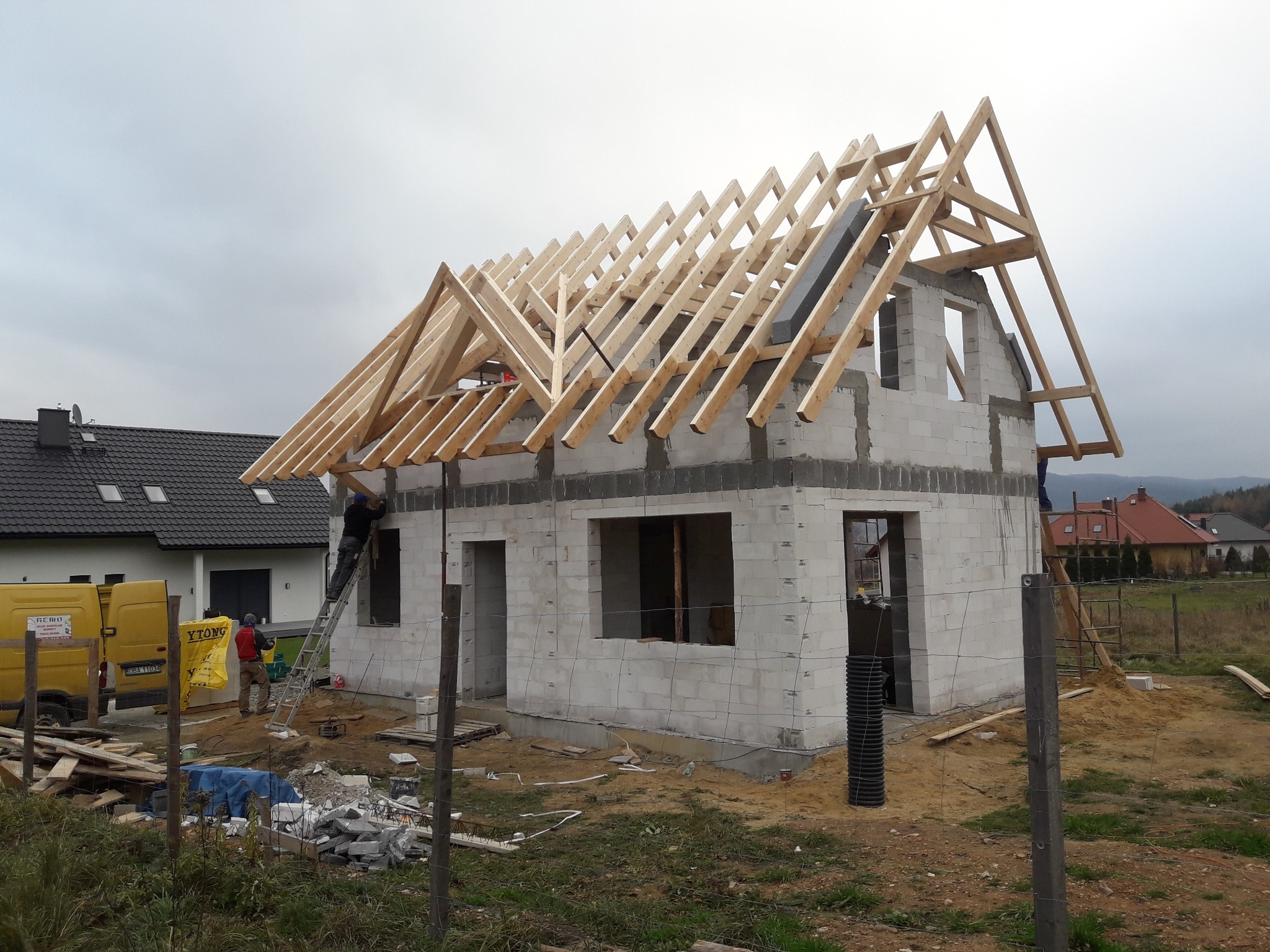 Budowa domu jednorodzinnego w trakcie wznoszenia konstrukcji dachu z drewnianych krokwi, ściany z bloczków betonowych, widoczny samochód dostawczy firmy budowlanej, pracownicy na drabinie...