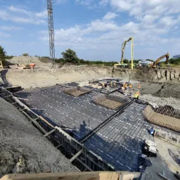 Przygotowanie terenu pod budynek: głęboki wykop z ułożoną folią, zbrojeniem i szalunkiem, na górze ekipa budowlana, betoniarka i koparka, w tle budynek biurowy.