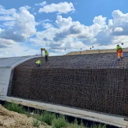 Trzech pracowników w odblaskowych ubraniach montuje zbrojenie na betonowej konstrukcji w kształcie tunelu, widok na rozległe niebo z chmurami.