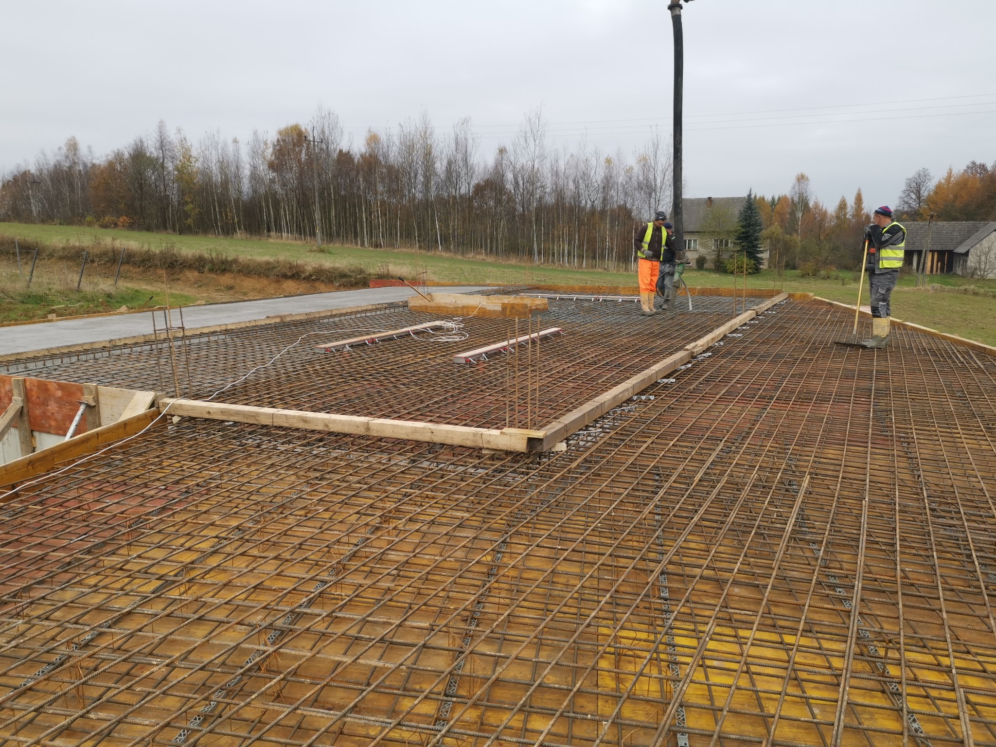 Proces wylewania betonu na przygotowane zbrojenie fundamentu, widoczni dwaj pracownicy w odblaskowych kamizelkach, jeden obsługuje pompę do betonu, drugi wyrównuje powierzchnię, w tle zabudowania...