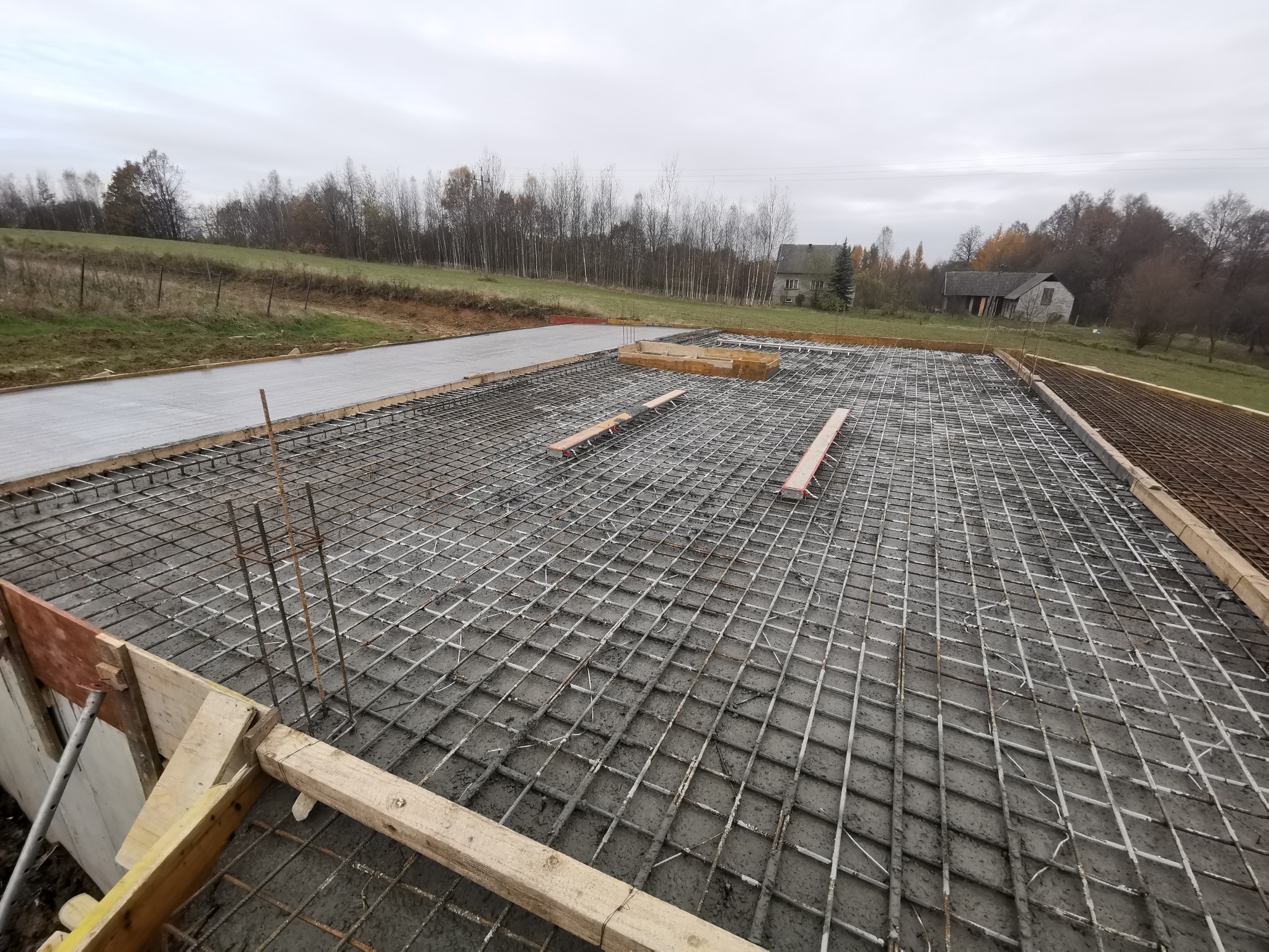 Siatka zbrojeniowa zalana betonem, przygotowana do wylania płyty fundamentowej, widok z góry na tle pochmurnego nieba i zabudowań gospodarczych.