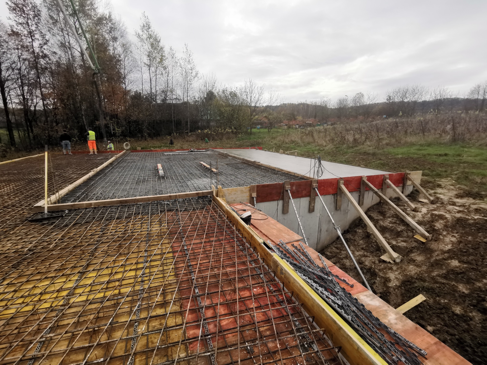 Zbrojenie fundamentów budynku z częściowo wylanym betonem, widoczna pompa do betonu i pracownicy w oddali, teren budowy w jesiennej scenerii.