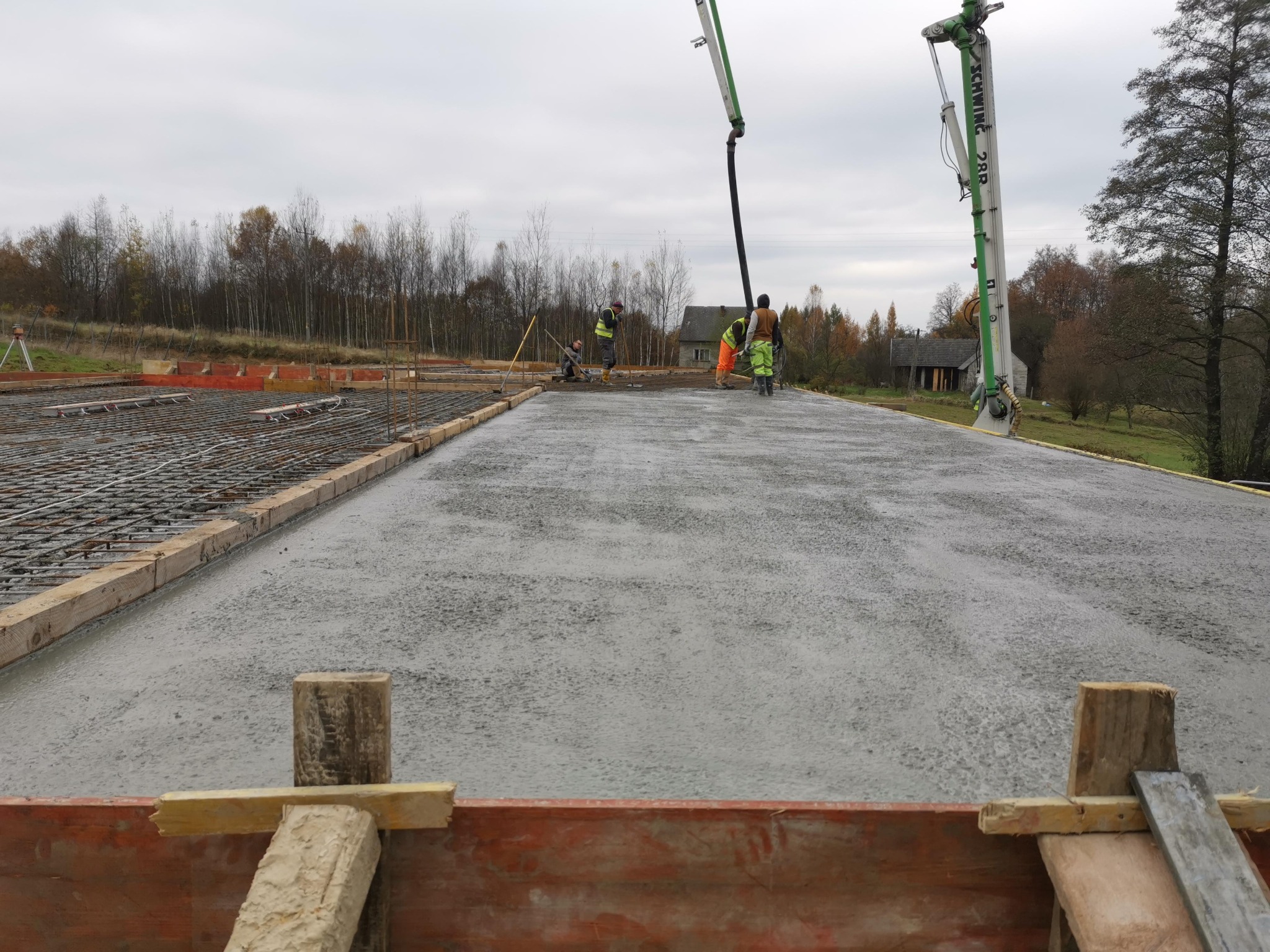 Wylewanie betonu na zbrojone fundamenty z użyciem pompy do betonu Schwing 28R, w tle pracownicy w odblaskowych kamizelkach i drzewa.