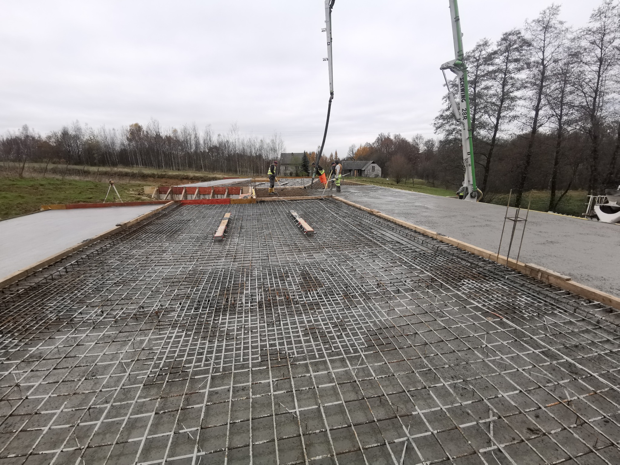 Wylewanie betonu z pompy na zbrojenie fundamentu, widoczni pracownicy w odblaskowych kamizelkach i drewniane szalunki, w tle drzewa i pochmurne niebo.