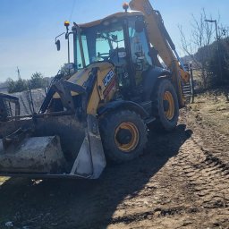 K.N-TURBO TRANS-KOP Kamil Napiórkowski - Żółta koparko-ładowarka JCB na nierównym, błotnistym terenie w słoneczny dzień. Widoczne ślady opon i elementy krajobrazu w tle. Maszyna gotowa do pracy.