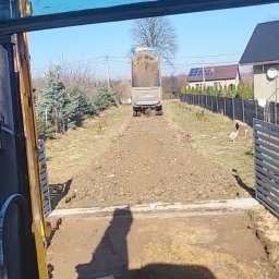 K.N-TURBO TRANS-KOP Kamil Napiórkowski - Widok z kabiny koparki na wywrotkę wysypującą ziemię na teren działki. W tle dom z panelami słonecznymi i drzewa bez liści. W kadrze fragment łyżki koparki.
