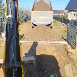 K.N-TURBO TRANS-KOP Kamil Napiórkowski - Żółta koparka i wywrotka z ładunkiem ziemi na placu budowy. Widok z kabiny koparki, błękitne niebo w tle. Prace ziemne w toku, teren przygotowywany pod inwestycję.