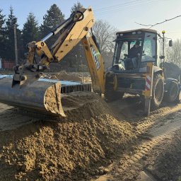 K.N-TURBO TRANS-KOP Kamil Napiórkowski - Żółta koparko-ładowarka JCB nabiera piasek z pryzmy, w tle wykop z zabezpieczeniem ścianek. Operator w kabinie. Słoneczny dzień.
