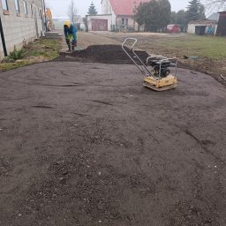 K.N-TURBO TRANS-KOP Kamil Napiórkowski - Przygotowanie podłoża: ubijarka wibrująca na wyrównanym terenie, pracownik z łopatą w tle, budynek w stanie surowym. Prace ziemne na działce.