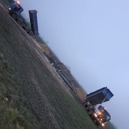 K.N-TURBO TRANS-KOP Kamil Napiórkowski - Dwa wywrotki w trakcie rozładunku na polu w pochmurny dzień. Widoczne pryzmy materiału i elementy konstrukcyjne. Ujęcie z boku, lekko pod kątem.