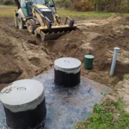Żółta koparko-ładowarka w wykopie ziemnym, obok dwa betonowe kręgi szamba z pokrywami, widoczne także rury kanalizacyjne i zielony pojemnik.