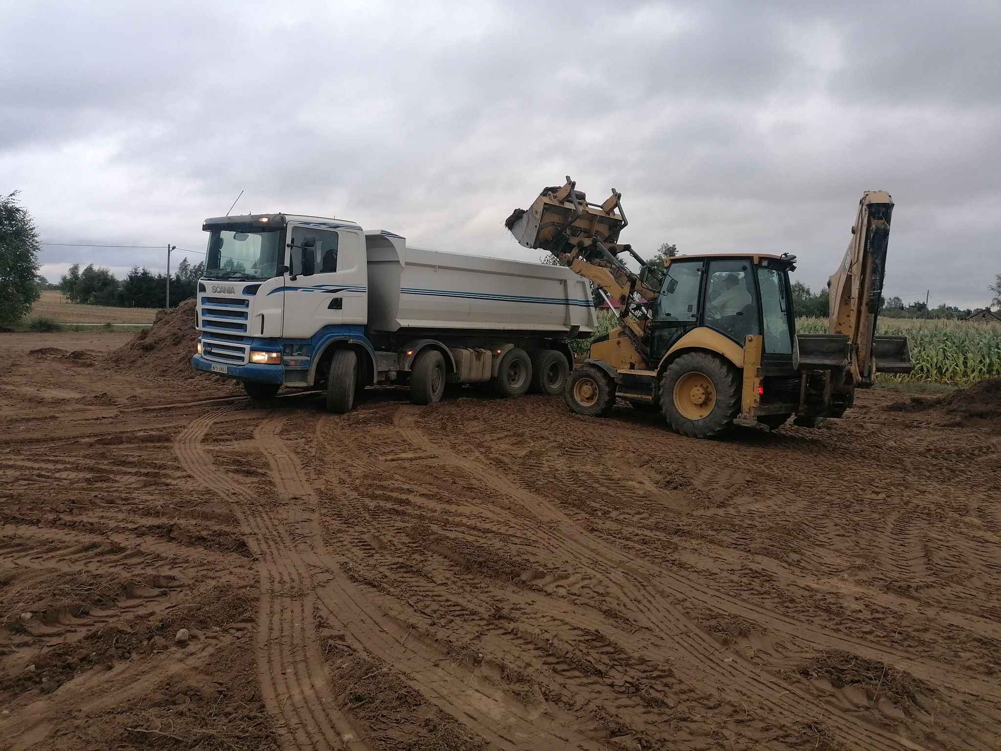 Żółta koparko-ładowarka nabiera ziemię do wywrotki Scania na placu budowy, widoczne ślady opon na ziemi, pochmurne niebo.