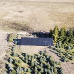 Widok z drona na instalację paneli słonecznych na gruncie, otoczoną drzewami iglastymi i polami. Widoczny pracownik przy instalacji.