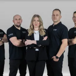 Zespół w czarnych koszulkach polo z logo firmy, stojący na tle szarego studia. Kobieta w garniturze pośrodku. Profesjonalny wizerunek firmy.