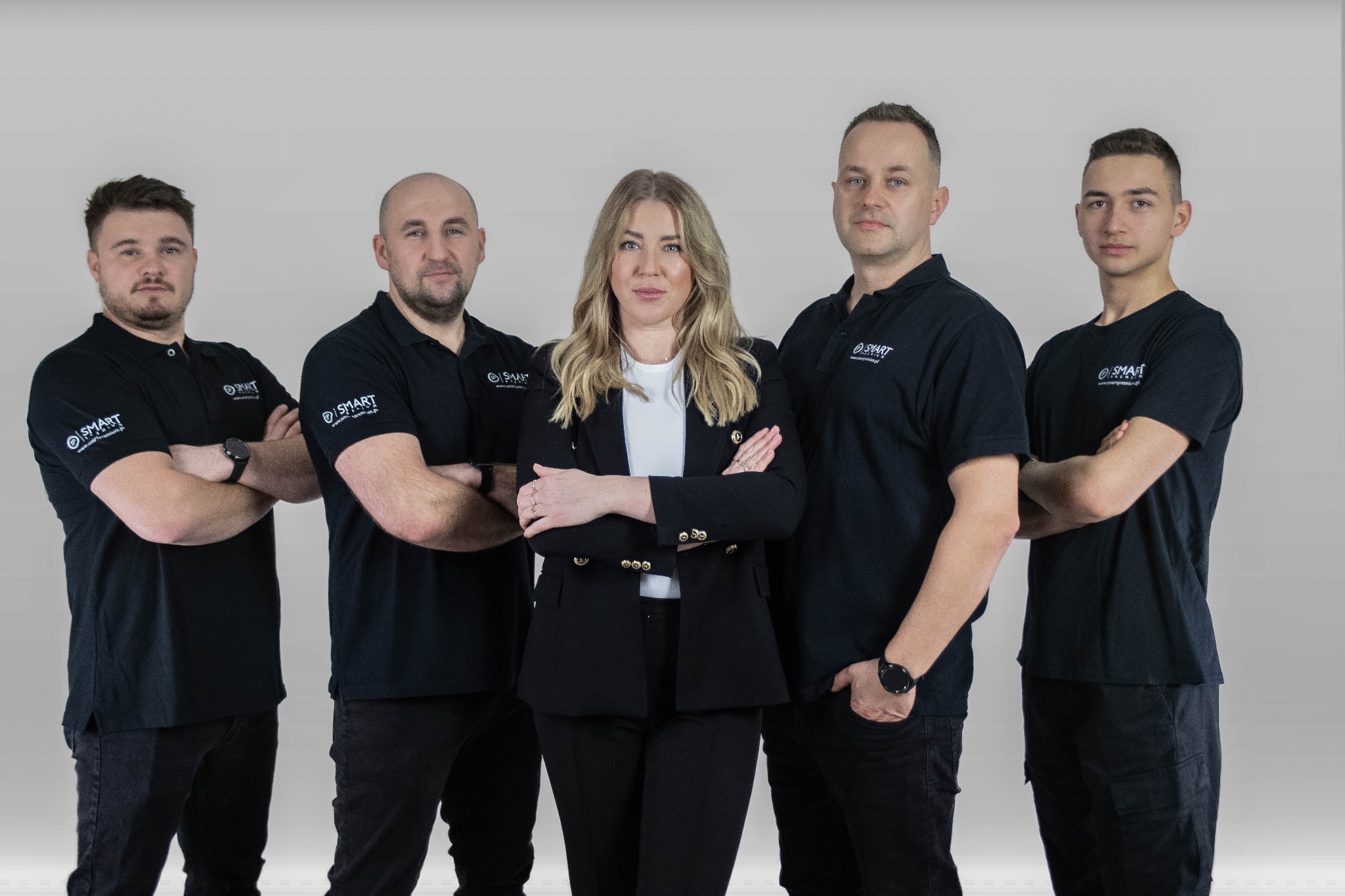 Zespół w czarnych koszulkach polo z logo firmy, stojący na tle szarego studia. Kobieta w garniturze pośrodku. Profesjonalny wizerunek firmy.