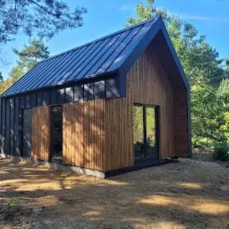 Nowoczesny dom o prostej bryle z drewnianą elewacją i ciemnym dachem z blachy na leśnej działce, widok z niskiej perspektywy.