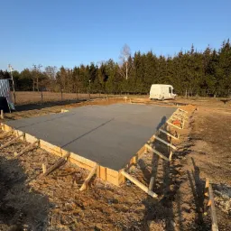 Wylany fundament pod budynek, z drewnianym szalunkiem, na działce z widocznym samochodem dostawczym w tle.