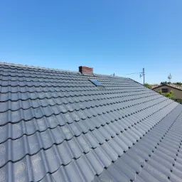 Szary dach z blachodachówki modułowej, widoczny komin z cegły i okno dachowe, błękitne niebo w tle.