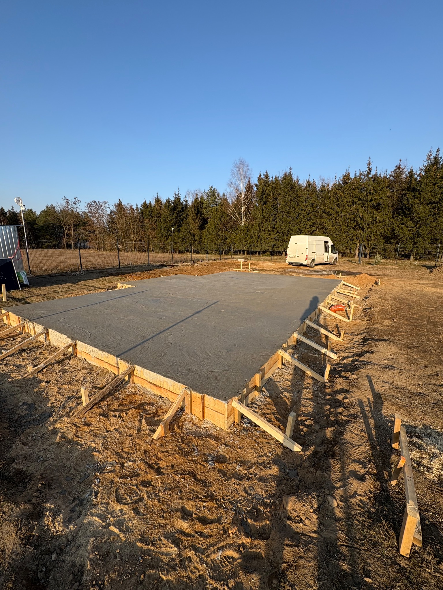 Wylany fundament pod budynek, z drewnianym szalunkiem, na działce z widocznym samochodem dostawczym w tle.