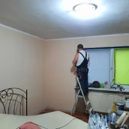 Mężczyzna w kombinezonie roboczym stoi na drabinie i wygładza ścianę w pokoju z łóżkiem, widoczne narzędzia i akcesoria malarskie na parapecie okna.