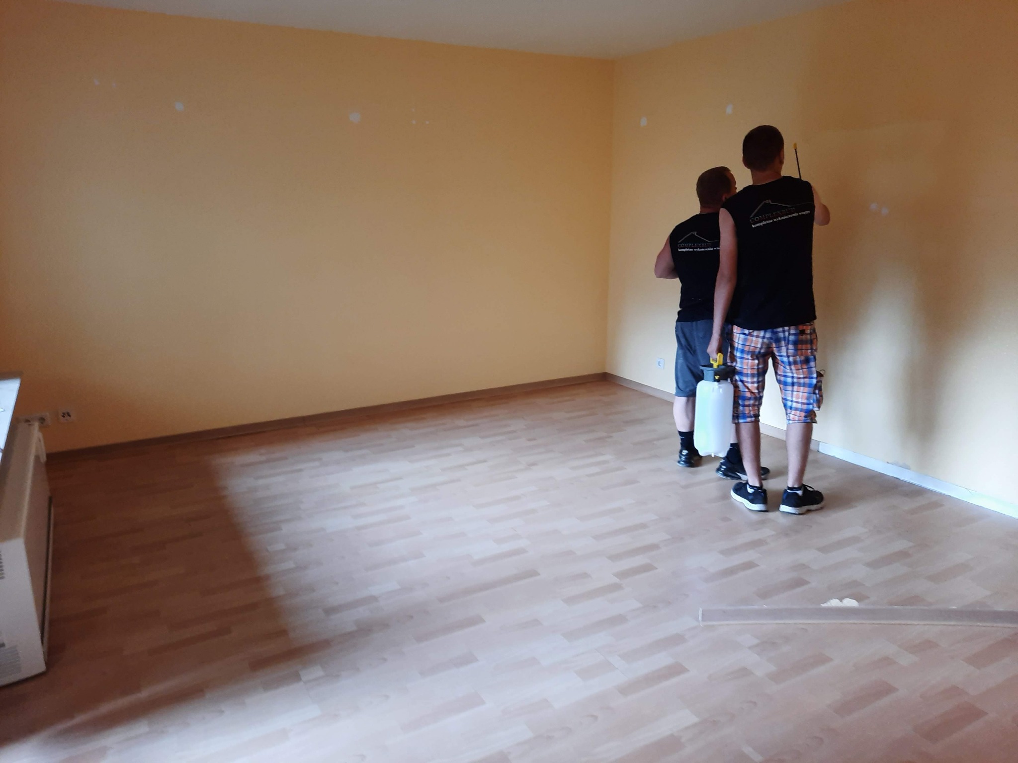 Dwóch mężczyzn w koszulkach z logo firmy maluje jasną ścianę w pustym pokoju z ułożonymi panelami podłogowymi; widoczny spryskiwacz i poziomica.
