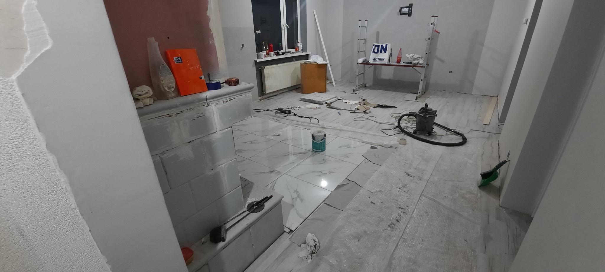 Wnętrze w trakcie prac wykończeniowych: świeżo położone błyszczące, białe płytki podłogowe imitujące marmur, widoczne narzędzia, odkurzacz przemysłowy, drabina i materiały budowlane w tle, ściany...