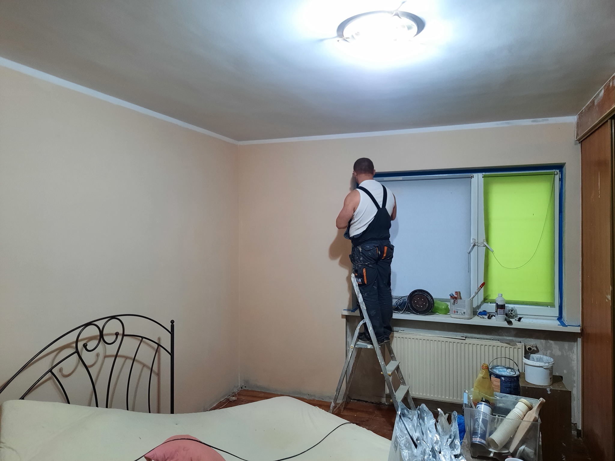 Mężczyzna w kombinezonie roboczym stoi na drabinie i wygładza ścianę w pokoju z łóżkiem, widoczne narzędzia i akcesoria malarskie na parapecie okna.