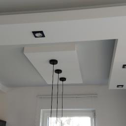 NowaMal Krzysztof Nowakowski Wykończenia Remonty Stolarstwo - Nowoczesny sufit podwieszany z oświetleniem punktowym, ceglana ściana w tle, minimalistyczna lampa wisząca z trzema czarnymi kloszami.