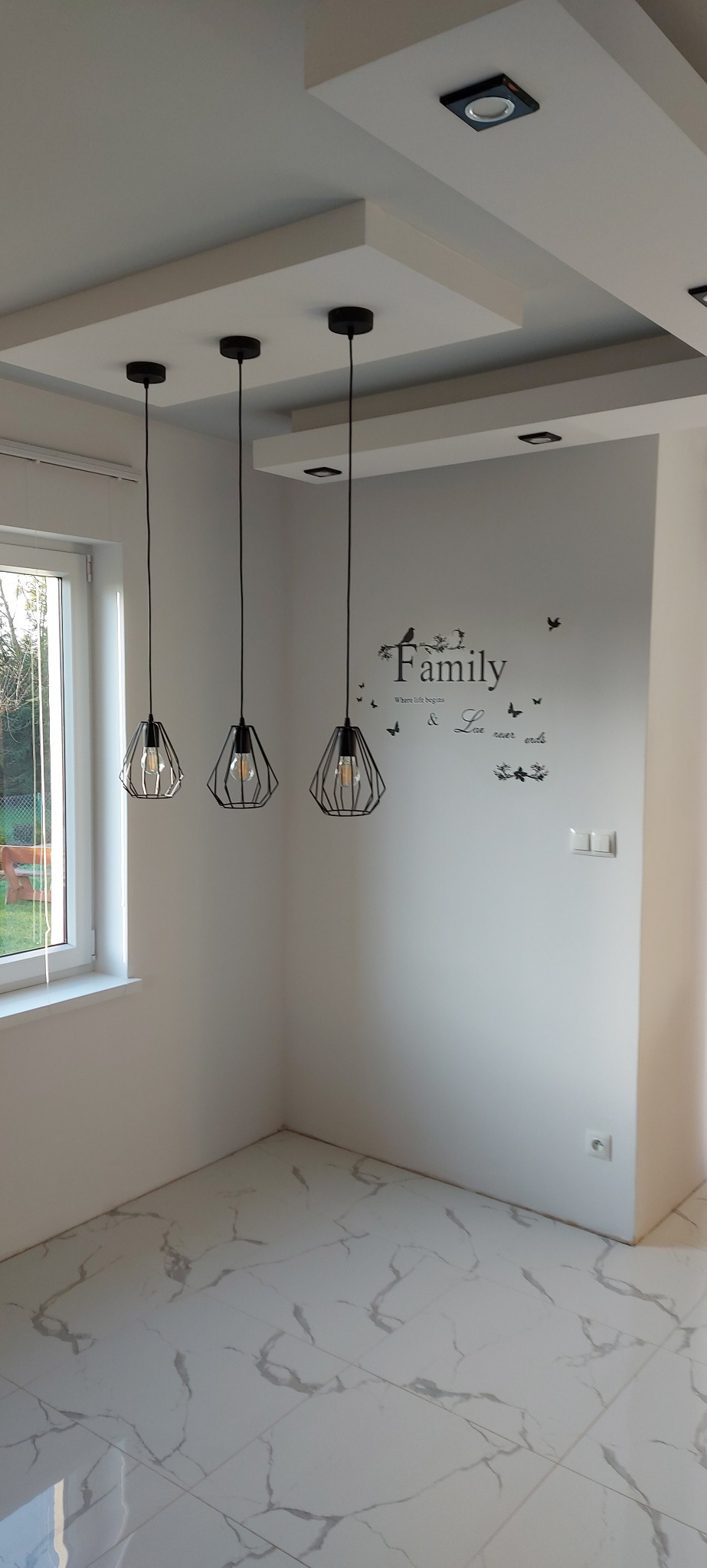 Wnętrze pokoju po remoncie: białe ściany z naklejką 'Family', marmurowe płytki podłogowe, trzy czarne lampy wiszące o ażurowych kloszach i ozdobny sufit z oświetleniem punktowym.