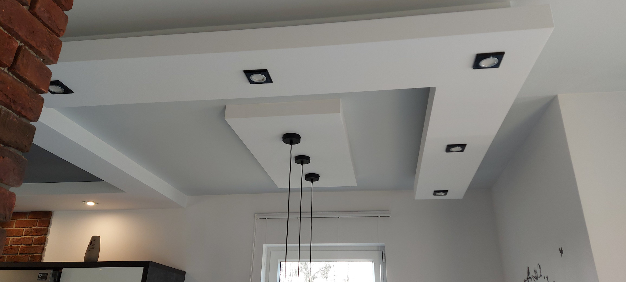 Nowoczesny sufit podwieszany z oświetleniem punktowym, ceglana ściana w tle, minimalistyczna lampa wisząca z trzema czarnymi kloszami.