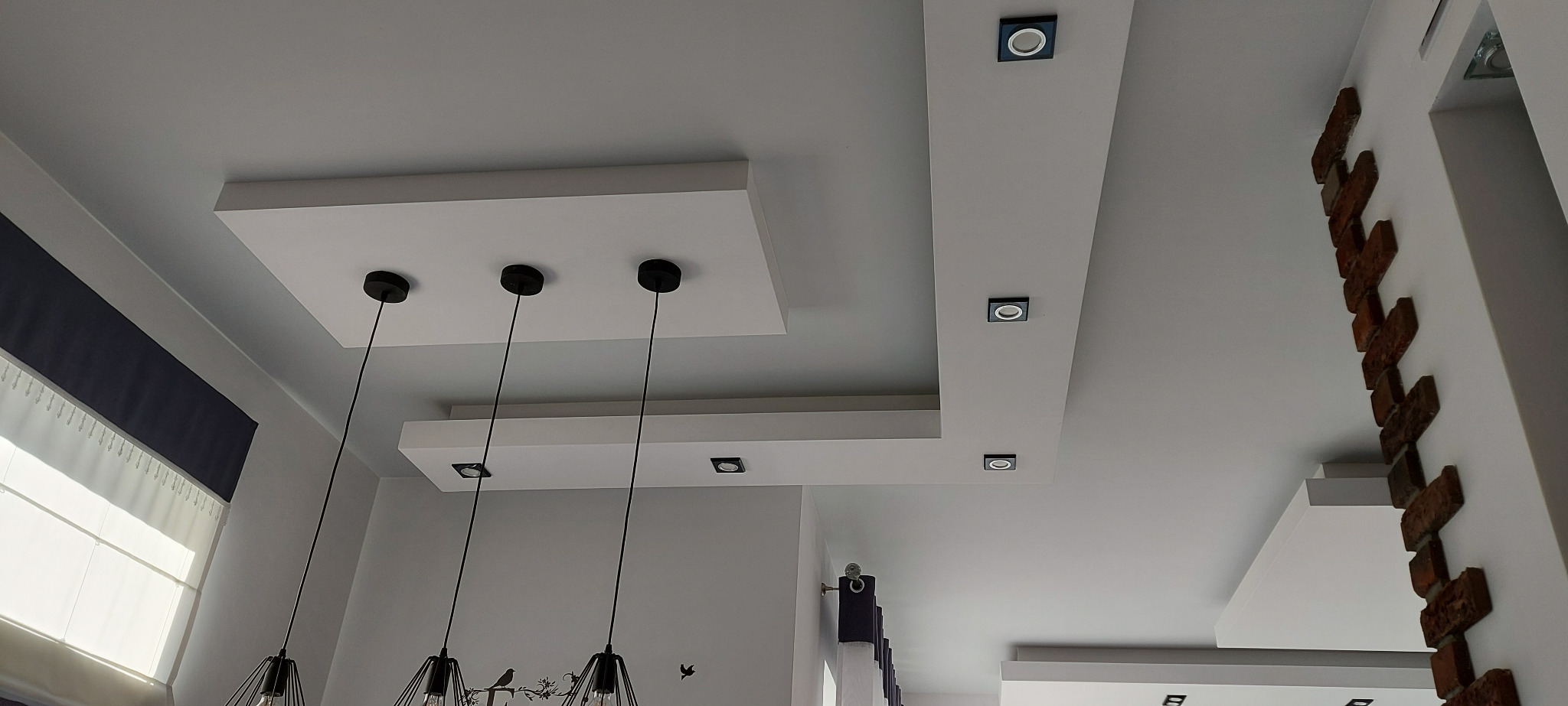 Wielopoziomowy sufit podwieszany z płyt kartonowo-gipsowych z oświetleniem punktowym i wiszącymi lampami, dekoracyjna ściana z cegły.