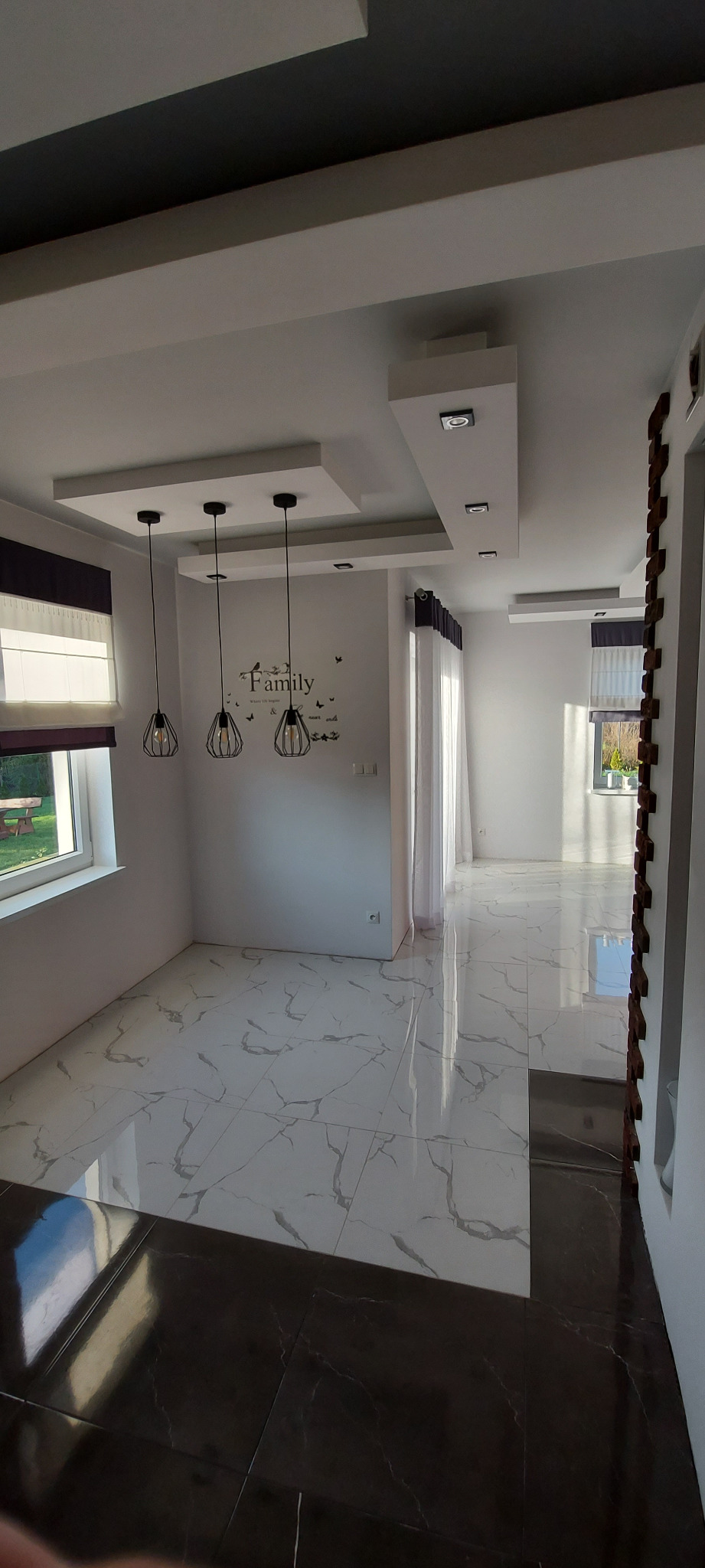 Wykończone wnętrze salonu z marmurową podłogą, podwieszanym sufitem z oświetleniem punktowym i trzema lampami wiszącymi nad ścianą z naklejką 'Family'.