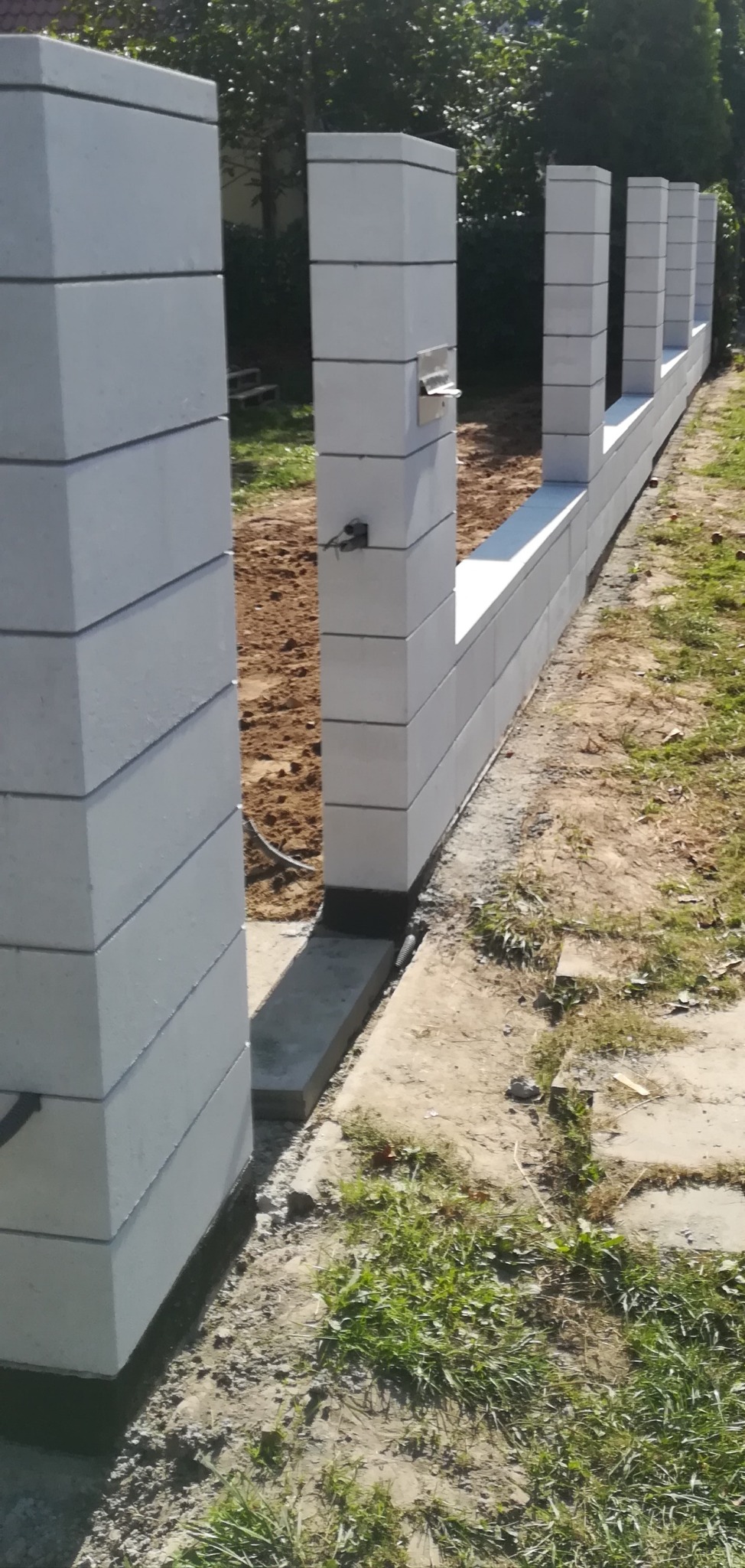 Wznoszenie ogrodzenia z białych, betonowych bloczków modułowych; widoczne słupki i podmurówka w trakcie budowy, z ziemią wokół.
