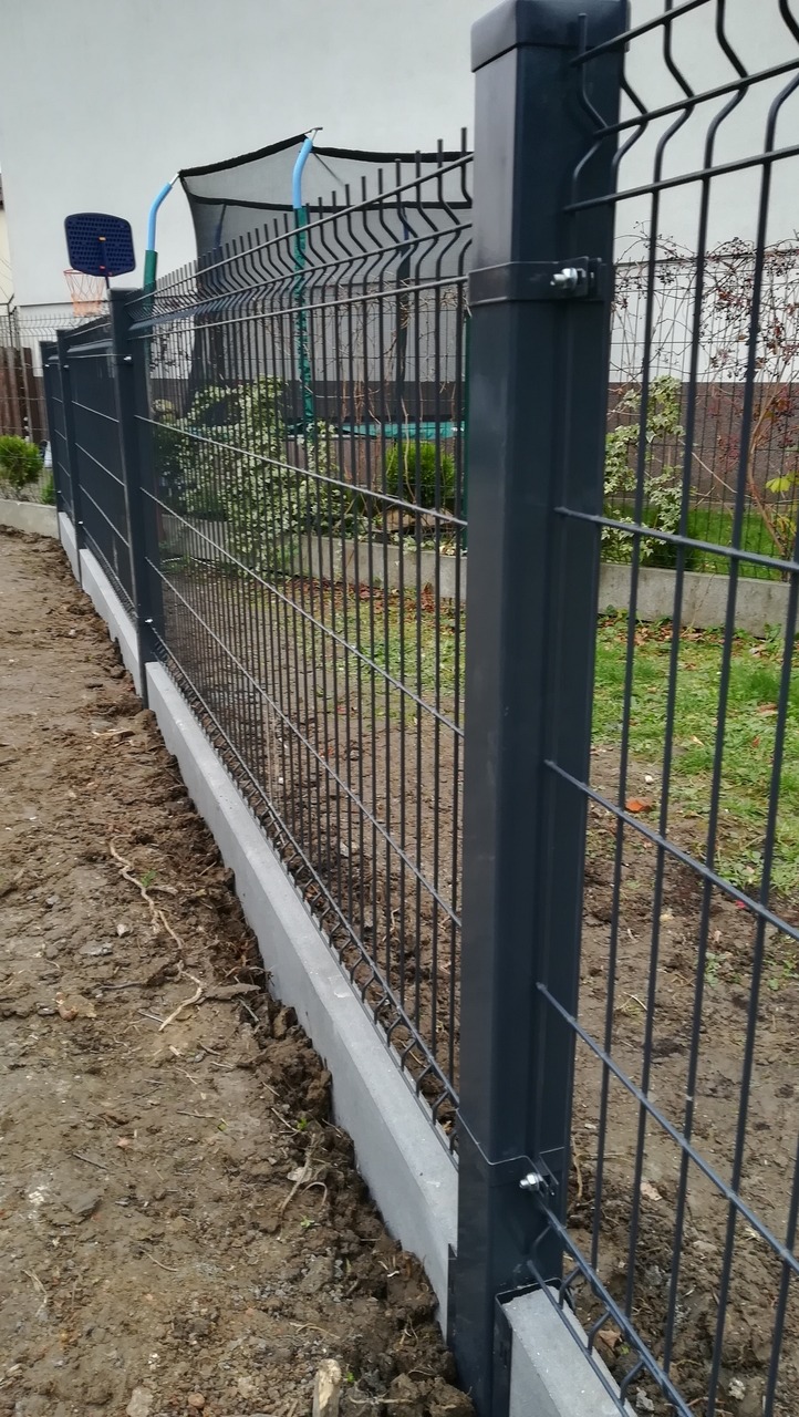 Panelowe ogrodzenie w kolorze grafitowym z betonową podmurówką, widok z bliska na słupki i siatkę, w tle trampolina i kosz do koszykówki.