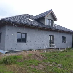 Dom jednorodzinny w trakcie ocieplania ścian zewnętrznych, widoczne szare płyty izolacyjne z charakterystycznymi otworami, czarne ramy okienne, metalowa drabina oparta o ścianę, teren wokół domu...